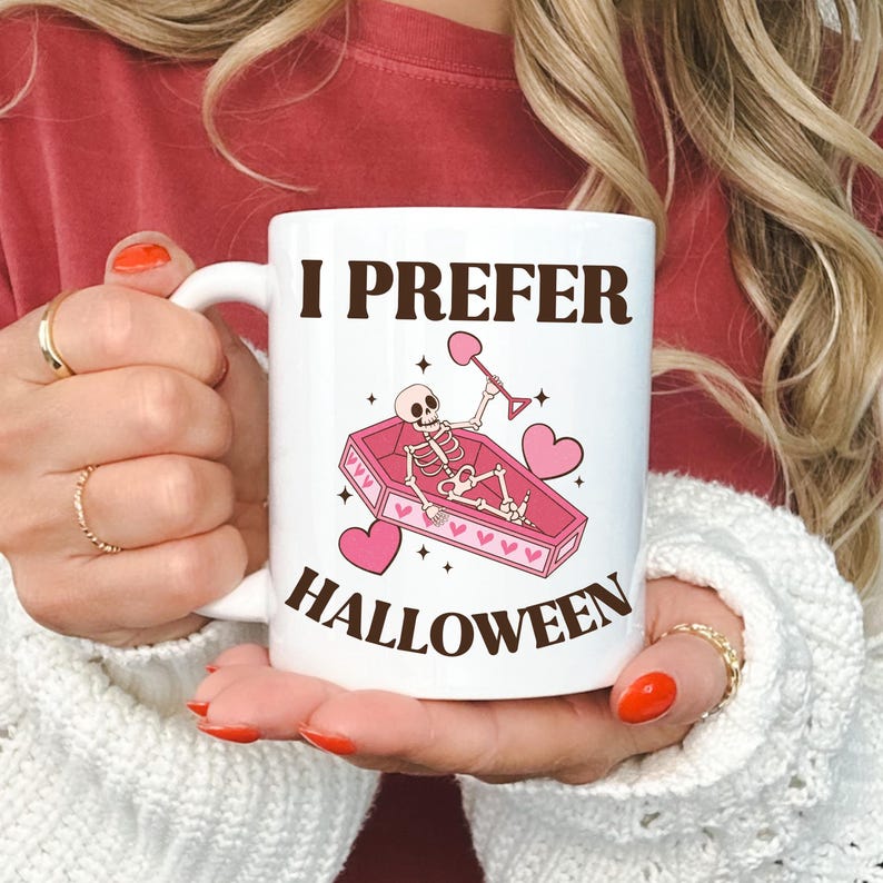 May include: White ceramic coffee mug with a pink and brown illustration of a skeleton in a coffin holding a shovel. The text "I prefer Halloween" is printed on the mug.