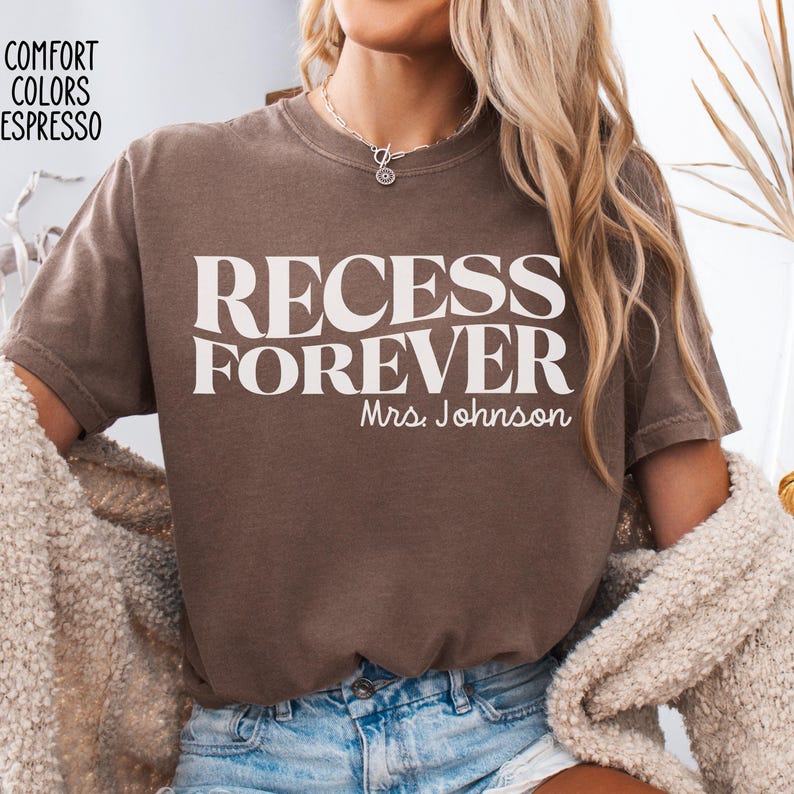 Puede incluir: Camiseta color espresso con las palabras "RECESS FOREVER" en letras blancas grandes. Debajo, "Mrs. Johnson" est&aacute; escrito en una fuente m&aacute;s peque&ntilde;a. La camiseta es parte de la colecci&oacute;n Comfort Colors.