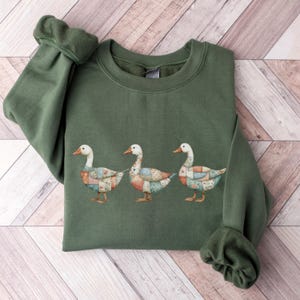 May include: An olive green sweatshirt featuring a design of three geese with patchwork-style bodies. The geese have white heads and orange beaks and feet. The sweatshirt has a crew neck and long sleeves.