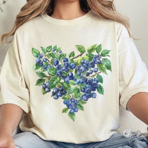 May include: A cream-colored t-shirt with a watercolor illustration of blueberries in a heart shape. The blueberries are a deep blue color and the leaves are green.