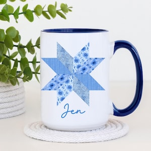 May include: White ceramic mug with a blue interior and handle. The mug features a blue star design with floral patterns and the name "Jen" in a script font. The mug sits on a white coaster, with greenery in the background.