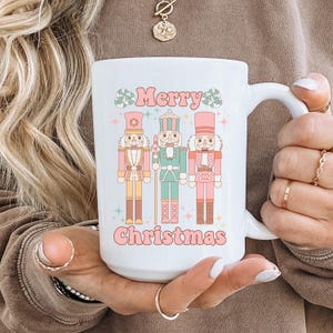 May include: White ceramic mug with a pink, green, and brown illustration of three nutcracker figurines. The mug has the text "Merry Christmas" in pink letters.