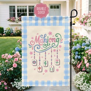 May include: A double-sided garden flag with a light beige background and a blue gingham border. The flag features the word "Mahjong" in pink and green script, along with hanging tiles and floral accents.
