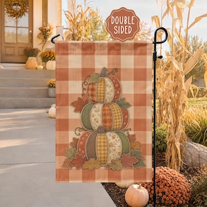 May include: A rectangular garden flag featuring a light orange and white buffalo check pattern. The center displays a stack of three pumpkins made of various fabric patches in orange, green, and yellow tones, surrounded by fabric autumn leaves.