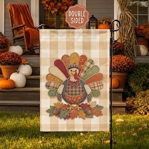 May include: A rectangular garden flag featuring a beige and white buffalo check pattern. The center displays a turkey with a colorful, patterned fan tail, wearing a plaid cap, standing on a pile of autumn leaves and acorns. Text reads DOUBLE SIDED.