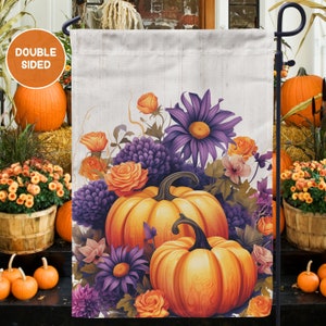 May include: A double-sided garden flag with a fall theme. The flag features two pumpkins, one on top of the other, surrounded by purple and orange flowers. The background is a whitewashed wood.