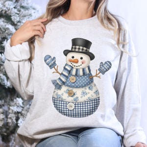 May include: A light gray sweatshirt featuring a whimsical snowman design. The snowman has a black top hat, a carrot nose, and a blue and white patterned scarf and mittens. The snowman's body is adorned with floral and gingham patterns.