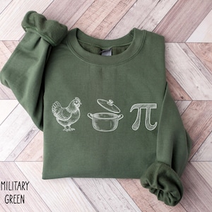 May include: Military green sweatshirt with a white graphic of a chicken, a pot, and the pi symbol. The text "MILITARY GREEN" is printed in white on the bottom left of the sweatshirt. The sweatshirt has a crew neck and long sleeves.