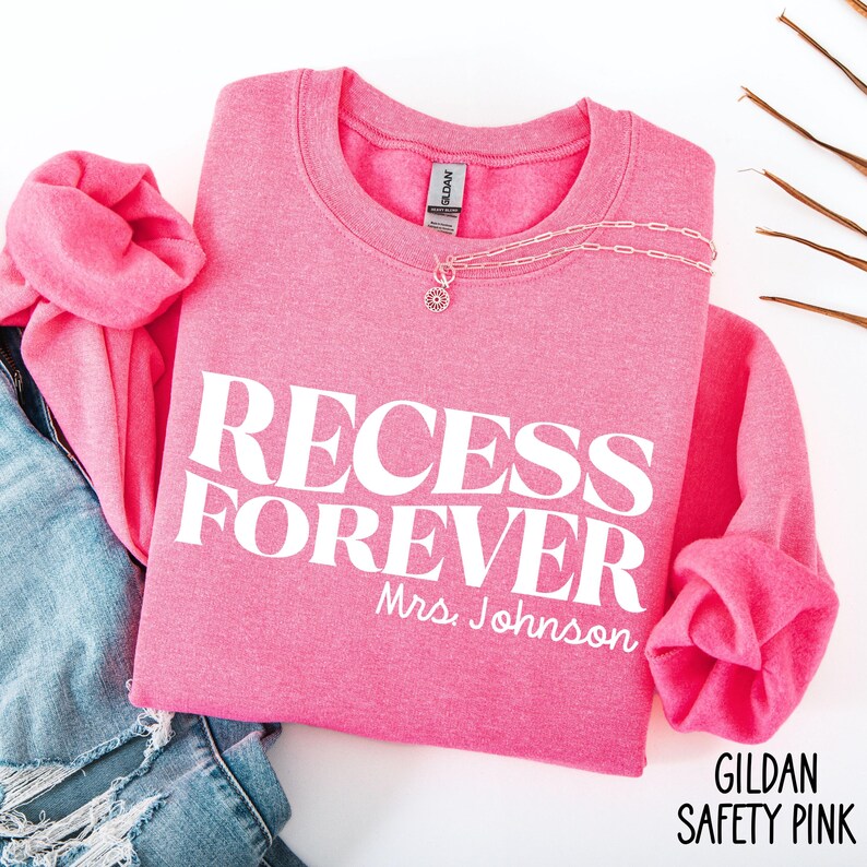 Puede incluir: Una sudadera rosa con las palabras "RECESS FOREVER" impresas en blanco. El nombre "Mrs. Johnson" est&aacute; debajo del texto. Un collar plateado con un colgante est&aacute; sobre la camisa. La sudadera est&aacute; doblada sobre unos vaqueros.