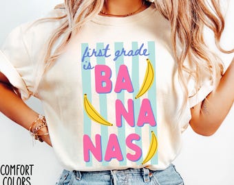 May include: Ivory-colored t-shirt with the text "first grade is BANANAS" in blue and pink, with yellow banana illustrations. The shirt also has the words "COMFORT COLORS IVORY" printed on the bottom left.