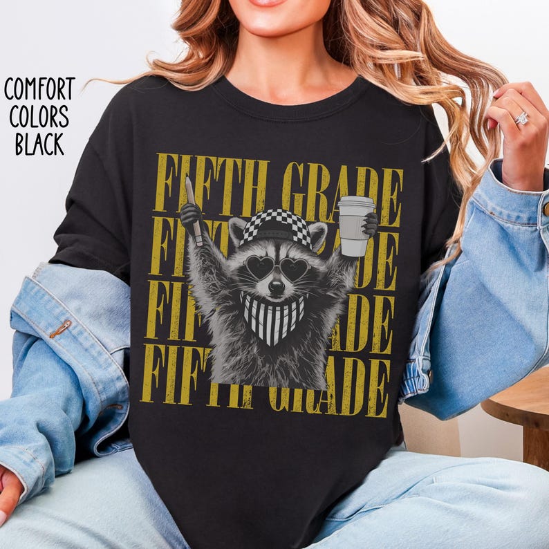May include: A black t-shirt featuring a graphic of a raccoon wearing a checkered cap, heart-shaped sunglasses, and a striped bandana. The raccoon holds a pencil and a coffee cup. The text FIFTH GRADE is repeated in gold behind the raccoon.
