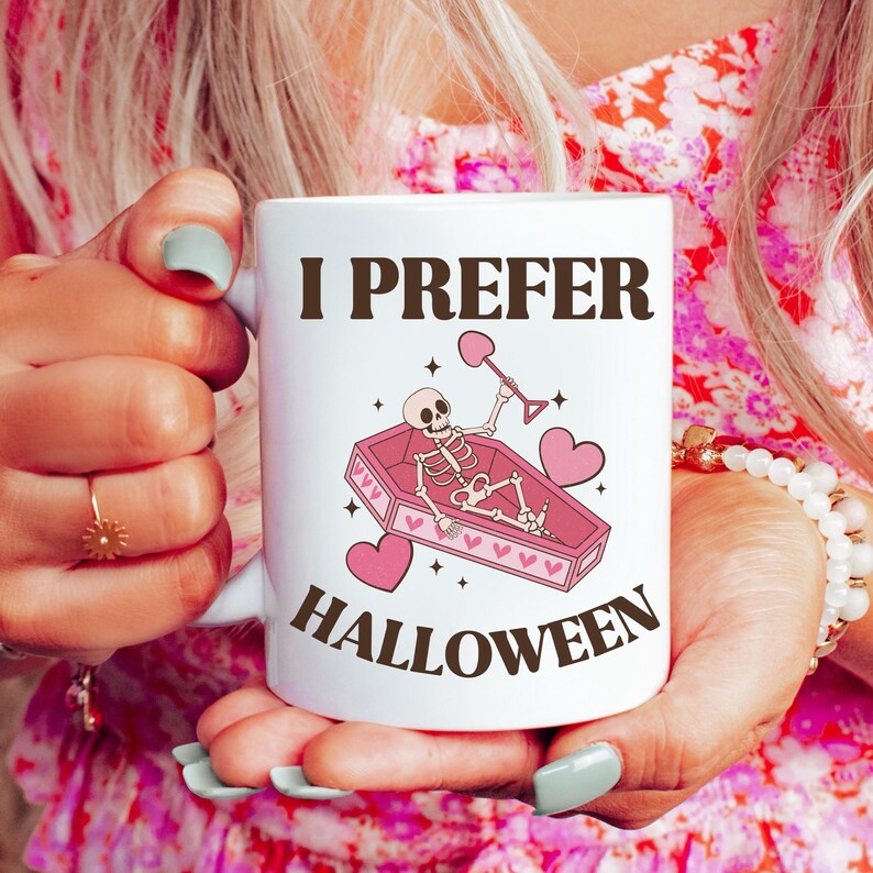 May include: White ceramic mug with a pink and black illustration of a skeleton in a coffin with hearts and the text "I prefer Halloween".