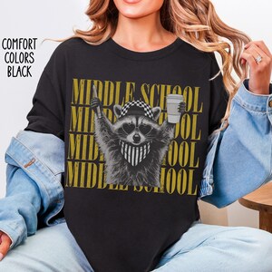 May include: A black t-shirt featuring a graphic of a raccoon wearing a checkered cap, heart-shaped sunglasses, and a striped bandana. The raccoon holds a pencil and a coffee cup. The text "MIDDLE SCHOOL" is repeated in gold behind the raccoon.