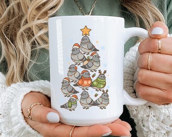 May include: White ceramic mug featuring a Christmas tree design made of cartoon pigeons wearing festive hats and sweaters. The tree is topped with a gold star and surrounded by snowflakes. The mug has a large handle.