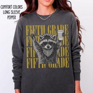 May include: A charcoal gray long-sleeve shirt featuring a graphic of a raccoon wearing a checkered cap, heart-shaped sunglasses, and a bandana. The raccoon holds a pencil and a coffee cup. The text FIFTH GRADE is repeated in gold behind the raccoon.