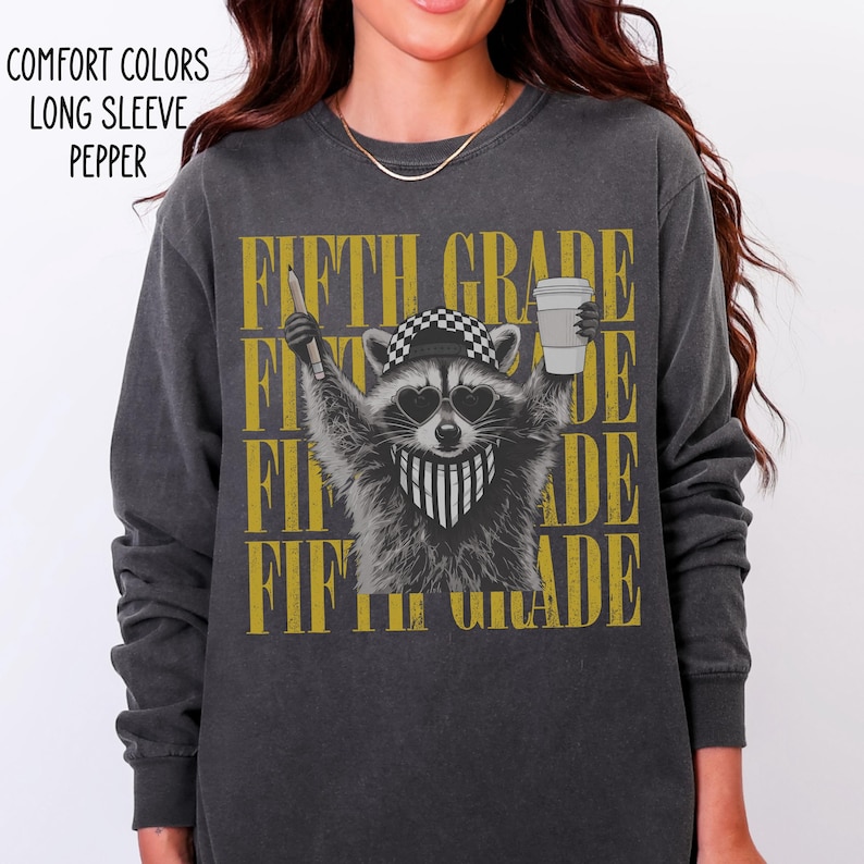 May include: A charcoal gray long-sleeve shirt featuring a graphic of a raccoon wearing a checkered cap, heart-shaped sunglasses, and a bandana. The raccoon holds a pencil and a coffee cup. The text FIFTH GRADE is repeated in gold behind the raccoon.