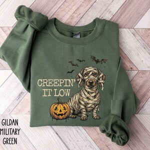 May include: An olive green crewneck sweatshirt featuring a graphic of a dachshund wrapped in mummy bandages sitting next to an orange jack-o'-lantern. Above the dog, the text reads "CREEPIN' IT LOW" with small black bats flying overhead.