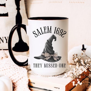 May include: White ceramic mug with a black handle and rim. The mug features the text "SALEM 1692" above a grayscale illustration of a witch's hat with a bow, and the phrase "THEY MISSED ONE" below the hat.