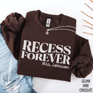Puede incluir: Sudadera marr&oacute;n chocolate oscuro con las palabras "RECESS FOREVER Mrs. Johnson" impresas en blanco. Un collar plateado con un colgante est&aacute; sobre la sudadera. La sudadera est&aacute; doblada junto a unos vaqueros azules.