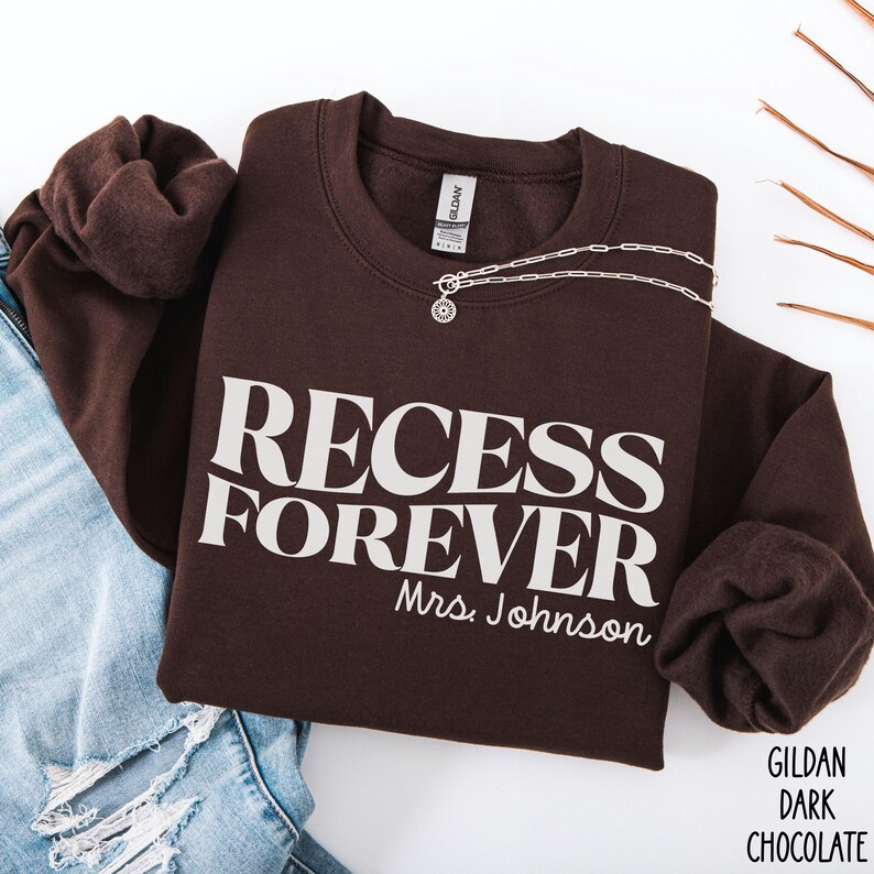Puede incluir: Sudadera marr&oacute;n chocolate oscuro con las palabras "RECESS FOREVER Mrs. Johnson" impresas en blanco. Un collar plateado con un colgante est&aacute; sobre la sudadera. La sudadera est&aacute; doblada junto a unos vaqueros azules.