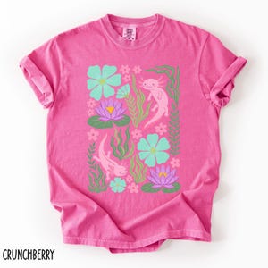 May include: A pink t-shirt featuring a colorful design of axolotls, flowers, and aquatic plants. The axolotls are pink, the flowers are teal and purple, and the plants are green. The word "CRUNCHBERRY" is printed at the bottom.