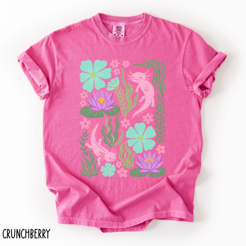 May include: A pink t-shirt featuring a colorful design of axolotls, flowers, and aquatic plants. The axolotls are pink, the flowers are teal and purple, and the plants are green. The word "CRUNCHBERRY" is printed at the bottom.