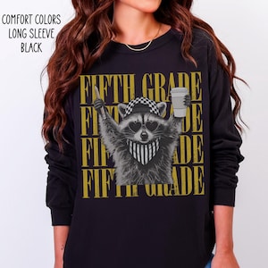 May include: A black long-sleeve shirt featuring a graphic of a raccoon wearing a checkered cap, heart-shaped sunglasses, and a striped bandana. The raccoon holds a pencil and a coffee cup. The text "FIFTH GRADE" repeats in gold behind the raccoon.