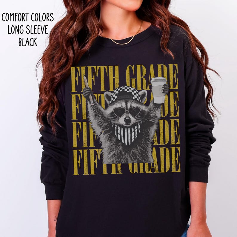 May include: A black long-sleeve shirt featuring a graphic of a raccoon wearing a checkered cap, heart-shaped sunglasses, and a striped bandana. The raccoon holds a pencil and a coffee cup. The text "FIFTH GRADE" repeats in gold behind the raccoon.