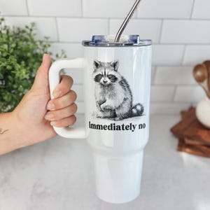 May include: White travel mug with a black and white illustration of a raccoon and the text "Immediately no." The mug has a white handle, a clear lid, and a silver straw.