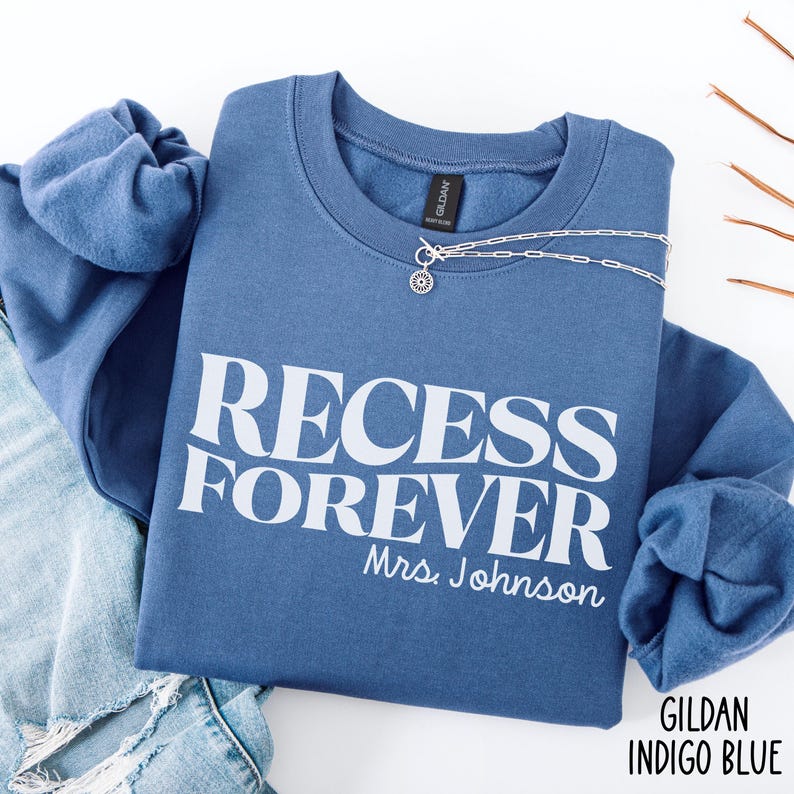 Puede incluir: Una sudadera azul con las palabras "RECESS FOREVER" en blanco, con "Mrs. Johnson" debajo. Un collar plateado con un colgante est&aacute; sobre la camisa. La sudadera est&aacute; doblada sobre un par de jeans.
