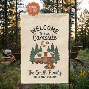 May include: A double-sided garden flag with the text "WELCOME to our Campsite" above a graphic of a camper, trees, and a campfire. The text "The Smith Family" and "PORTLAND, OREGON" are below the graphic. The flag is cream-colored and displayed outdoors.