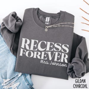 Puede incluir: Sudadera gris carb&oacute;n con las palabras "RECESS FOREVER" en letras blancas, con "Mrs. Johnson" debajo. Un collar plateado con un colgante est&aacute; colocado encima. La sudadera est&aacute; doblada sobre unos vaqueros azules.
