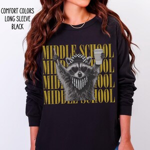 May include: A black long-sleeve shirt featuring a graphic of a raccoon wearing a checkered cap, heart-shaped sunglasses, and a bandana, holding a pencil and a coffee cup. The text "MIDDLE SCHOOL" is repeated in gold behind the raccoon.