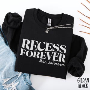 Puede incluir: Sudadera negra con el texto blanco "RECESS FOREVER Mrs. Johnson". Un collar plateado con un colgante est&aacute; colocado encima. La sudadera est&aacute; doblada sobre unos vaqueros azules.