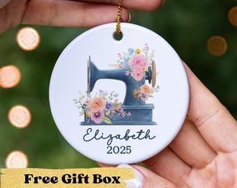 May include: A white ceramic ornament featuring a blue sewing machine design with floral accents. The ornament has the name "Elizabeth" and the year "2025" printed on it. A gold string is attached for hanging.