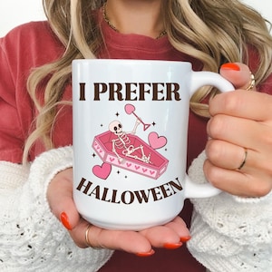 May include: White ceramic coffee mug with a black and pink design. The mug has a handle and says "I prefer Halloween" with a skeleton in a coffin and hearts.