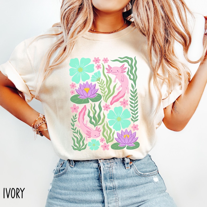 May include: Ivory-colored t-shirt featuring a colorful floral and axolotl design. The design includes pink axolotls, teal flowers, purple water lilies, and green foliage. The word "IVORY" is printed at the bottom left.
