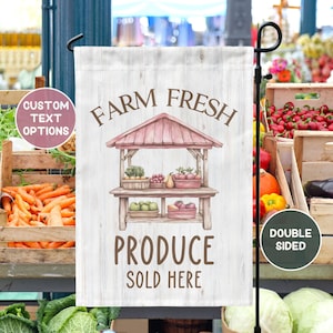 May include: A white wooden sign with the text "Farm Fresh Produce Sold Here" in brown lettering. The sign features a watercolor illustration of a red and white produce stand with fresh fruits and vegetables. The sign is double-sided and is perfect for a garden or farm.