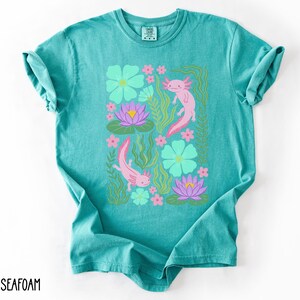 May include: Seafoam green t-shirt featuring a rectangular design with pink axolotls, teal flowers, purple water lilies, and green aquatic plants. The shirt has a crew neck and short sleeves. The word "SEAFOAM" is printed in black at the bottom left.