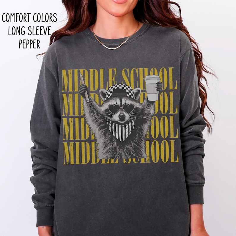 May include: A charcoal gray long-sleeve shirt featuring a graphic of a raccoon wearing a checkered cap, heart-shaped sunglasses, and a striped bandana. The raccoon holds a pencil and a cup. The text MIDDLE SCHOOL is repeated in gold behind it.
