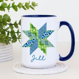 May include: White ceramic mug with a blue interior and handle. The mug features a star design in shades of green and blue, with the name "Jill" in a script font. The mug is sitting on a white coaster.