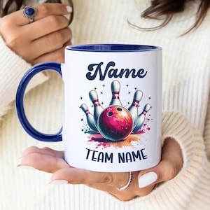 May include: White ceramic mug with a blue interior and handle. The mug features a bowling-themed design with a red and pink bowling ball and pins. The text "Name" and "Team Name" are displayed in a stylized font.