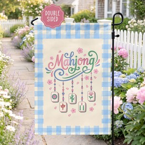 May include: A double-sided garden flag with a light beige background and a blue gingham border. The flag features the word "Mahjong" in pink and green script, along with hanging tiles and floral accents. The flag is displayed on a black metal stand.