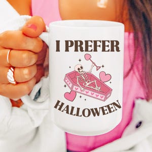 May include: White ceramic mug with a pink and brown design. The mug features a skeleton in a pink coffin with hearts and the text "I prefer Halloween".