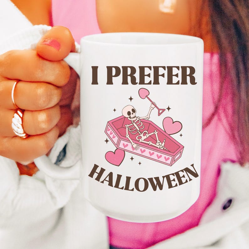 May include: White ceramic mug with a pink and brown design. The mug features a skeleton in a pink coffin with hearts and the text "I prefer Halloween".