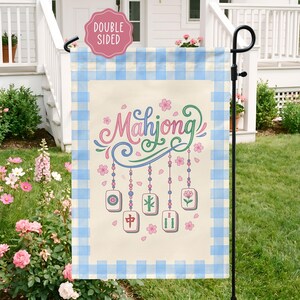 May include: A double-sided garden flag with a light beige background and a blue gingham border. The flag features the word "Mahjong" in pink and green script, along with hanging tiles and floral accents. The flag is displayed on a black metal stand.