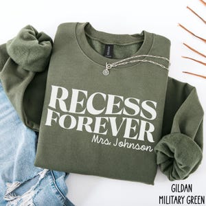 Puede incluir: Una sudadera verde oliva con las palabras "RECESS FOREVER" impresas en blanco, con "Mrs. Johnson" debajo. Un collar plateado con un colgante est&aacute; sobre la camisa. La esquina inferior derecha de la camisa tiene las palabras "GILDAN MILITARY GREEN".