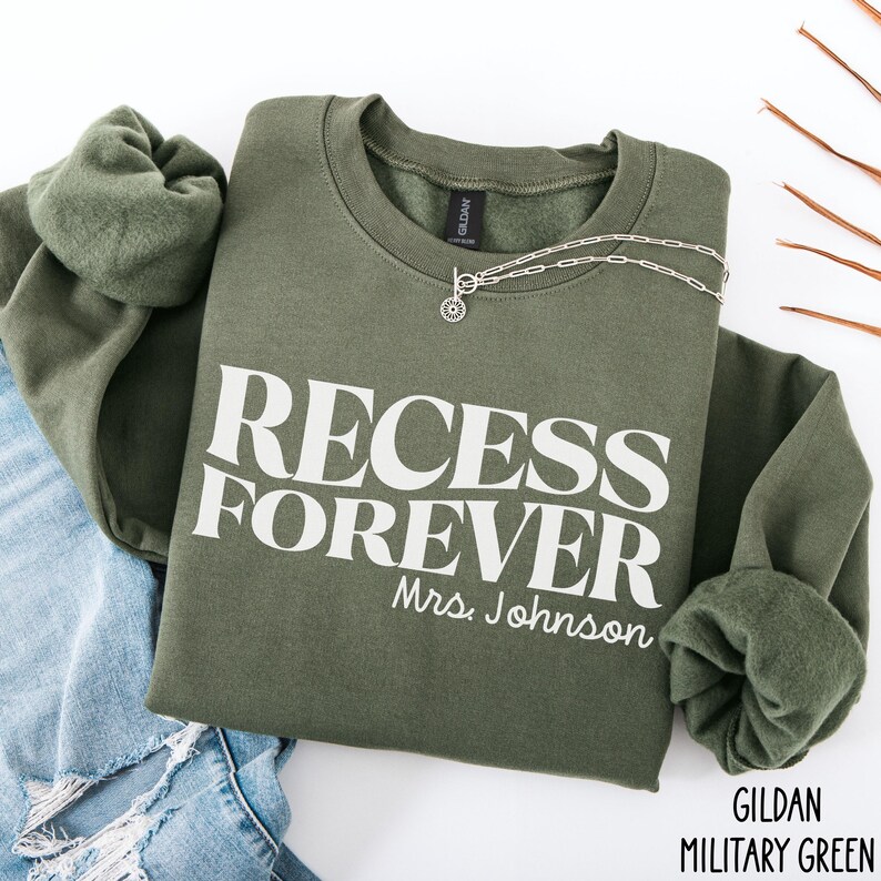 Puede incluir: Una sudadera verde oliva con las palabras "RECESS FOREVER" impresas en blanco, con "Mrs. Johnson" debajo. Un collar plateado con un colgante est&aacute; sobre la camisa. La esquina inferior derecha de la camisa tiene las palabras "GILDAN MILITARY GREEN".