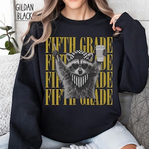 May include: A black long-sleeve sweatshirt featuring a graphic of a raccoon wearing a checkered cap, heart-shaped sunglasses, and a striped bandana. The raccoon holds a pencil and a coffee cup. The text FIFTH GRADE is repeated in yellow.