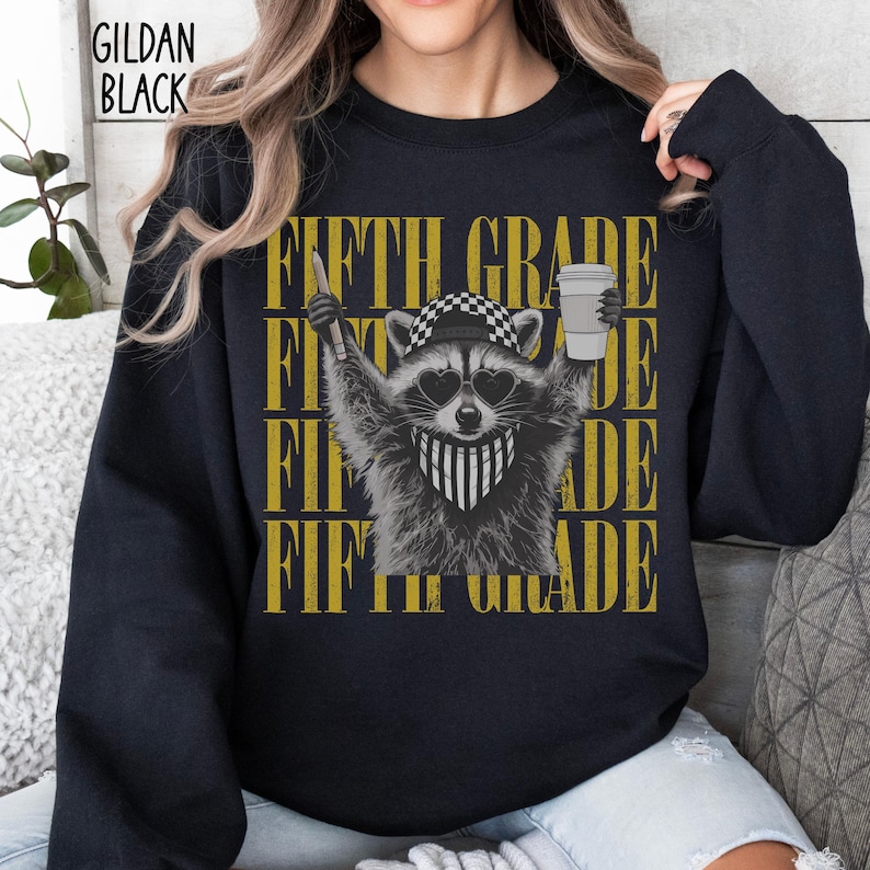 May include: A black long-sleeve sweatshirt featuring a graphic of a raccoon wearing a checkered cap, heart-shaped sunglasses, and a striped bandana. The raccoon holds a pencil and a coffee cup. The text FIFTH GRADE is repeated in yellow.